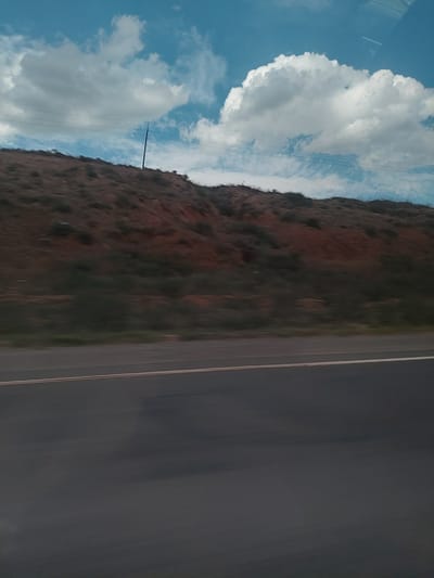 The image captures a scenic landscape from the perspective of a moving vehicle, with the view oriented as if looking out a side window. At the bottom right, a dark asphalt road is visible, showing blur consistent with motion. A faint white line marks the edge of the road, possibly indicating a shoulder.

To the left of the road, a large hillside or mountain rises, covered in sparse vegetation featuring muted shades of brown, green, and some reddish tones, suggesting arid or rocky terrain. The texture of the slope shows varied natural formations. Extending upwards from the hillside into the sky, a dark utility pole with several thin power lines is visible.

The upper portion of the image is dominated by a bright blue sky filled with large, fluffy white cumulus clouds, indicating fair weather during the daytime. Some faint reflections, possibly from a vehicle window frame, can be seen in the upper left corner of the sky. The overall scene suggests a journey through a rural or mountainous region, potentially in the area of Paruyr Sevak, Armenia, as indicated by the provided context. No text is visible in the image.