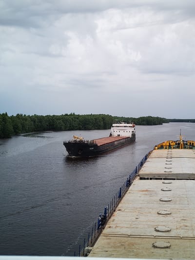The image shows a wide, dark river flowing through a dense, green forest. The sky is overcast with heavy, gray clouds. In the foreground, the deck of a large vessel is visible, extending towards the right side of the frame. It has a textured, metallic surface with several circular hatches. In the middle ground, a black freighter is sailing down the river, its bow pointed towards the viewer. This freighter has a white superstructure at its stern and appears to be carrying a cargo of bulk material on its deck, covered with a reddish-brown tarp. The freighter has the name "MEGAZH" visible on its hull. The riverbanks are lined with lush green trees, indicating a natural, possibly rural, environment. The overall scene is one of maritime activity on a waterway surrounded by nature, under a cloudy sky. The location is identified as Novokemskii, Russia.