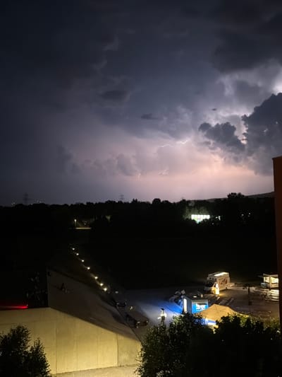 An outdoor scene at night in Glattbrugg, Switzerland, captures a dramatic thunderstorm with visible lightning strikes illuminating the dark, cloudy sky. The foreground features a concrete structure with a sloped surface, lined with small, glowing lights. Several trees are visible in the lower right and left corners of the frame. In the middle ground, a dark, tree-lined landscape stretches across the horizon. A single streetlight illuminates a path in the distance. Further into the scene, a small gathering appears to be taking place, with food trucks and tents set up. One white food truck with illuminated windows is prominently visible, as is a yellow tent. Several other illuminated structures and objects, possibly tables and chairs, are scattered around the area. High-voltage power lines and pylons are visible on the horizon to the left. The overall atmosphere is one of a stormy evening event.