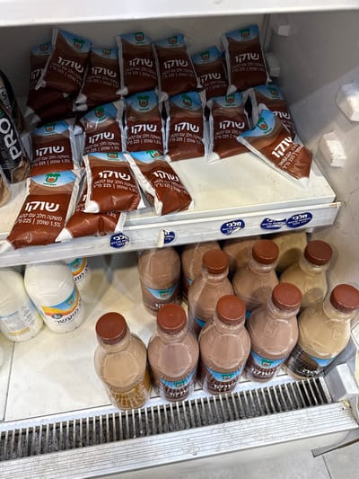 The image shows a refrigerated shelf in a convenience store in Kiryat Ekron, Israel (latitude: 31.865, longitude: 34.815). The top shelf is stocked with individually packaged chocolate milk cartons, all with the same Hebrew branding. The bottom shelf is stocked with nine chilled bottles of chocolate milk.  The bottom shelf's bottles have a different label than the cartons on the top shelf.  The shelves are white and metal.  The refrigerator is white and metal, with a gray floor.