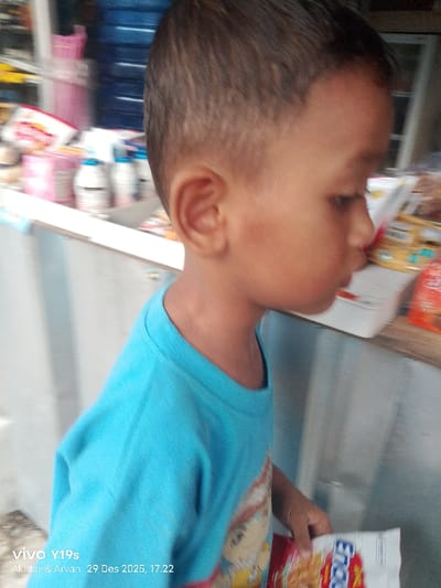 A young child is depicted in profile, facing right, standing within a retail establishment in Jagapura Kidul, Indonesia. The child, with short dark hair, wears a light blue t-shirt adorned with a partially visible graphic. The right hand holds a white and red snack package, partially marked "Enou," which appears to contain puffed food items. The blurred background reveals shelves or a counter stocked with various packaged goods, including pink boxes, white bottles, and additional snack packets. A large blue water dispenser is visible in the upper left background. The child appears to be looking downwards. The image is timestamped December 29, 2025, at 17:22.
