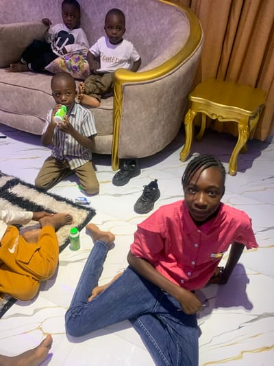 A high-angle, interior shot shows five children of varying ages. Three are seated on a large, gold-trimmed couch. The child on the right is wearing a striped shirt and holding a green and white bottle. The other two are in white shirts with dark pants. A fourth child, wearing a yellow sweatshirt, is partially visible on a black and white rug in the lower right corner of the image. A fifth person, who is wearing a pink shirt and blue jeans, is laying on the marble floor. Two pairs of black shoes, a small blue toy truck, and several green and white bottles are scattered on the floor. A gold accent table is next to the couch. The location appears to be a home in Dong, Nigeria.
