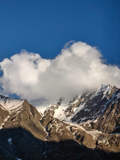 The image shows a mountain range under a bright blue sky with white clouds. The peaks of the mountains are covered in snow, with rocky slopes visible in between. Sunlight casts shadows on the mountains, highlighting the rugged terrain. The clouds are large and puffy, obscuring the tops of some of the peaks. The scene appears to be a remote, natural landscape. There are no people or man-made objects visible in the image. The location is described as Kead, India, but this cannot be verified from the image alone.