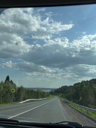 The image depicts a view from inside a vehicle traveling on a paved road near Ir'yak, Russia. The road curves gently to the right and has a white line dividing the lanes. A metal guardrail runs along the right side of the road, separating it from a forested area. A yellow and red traffic sign indicating a sharp curve is visible on the right side of the road. In the distance, beyond the trees, there appears to be a body of water. The sky is filled with numerous white and gray clouds. The viewpoint is from within a vehicle, evidenced by the vehicle frame on the left and bottom right of the image.