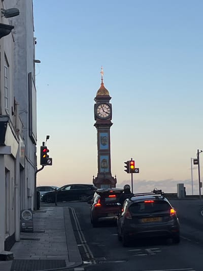 The image depicts the Weymouth Jubilee Clock Tower on the Esplanade in Weymouth, United Kingdom. The clock tower, a tall, ornate structure with a golden dome and a weather vane, stands centrally, displaying approximately 12:45 on its clock face. The scene is a street view under a clear, light blue sky with a faint warm glow near the horizon.
In the foreground, a two-lane asphalt road with double white lines shows vehicular traffic halted. A dark grey Ford Fiesta, license plate LJ65 UKD, equipped with a roof rack, is closest to the viewer. Immediately behind it is a red Volvo hatchback with a roof box. A person on a dark motorcycle, wearing a dark helmet and jacket, is situated behind the red car. Further back, a dark estate car is visible turning a corner. Two sets of traffic lights are present: one on the left, attached to a pole, and another on the right, both displaying illuminated red and amber signals. To the left, a light-colored building with a visible window and gutter line borders a paved sidewalk, which features a partially obscured sign for a coffee house. The background includes a distant horizon with subtle cloud formations.