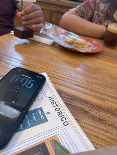 The image depicts a person seated at a wooden table in what appears to be a restaurant setting. The person's left hand and part of their arm are visible, wearing a patterned sleeve. On the table, there's a plate with food wrapped in plastic, a glass of liquid, a newspaper with the heading "HISTORICO" and the name "MAMÁ HIGO" labeled "RESTAURANTE". A black iPhone displays the time "6:05" and date "Sáb 2 de may" (Saturday, May 2). Based on the restaurant's name, "MAMÁ HIGO" on the newspaper, the location could be San Cristóbal, Mexico, as "MAMÁ HIGO" is a restaurant located there.