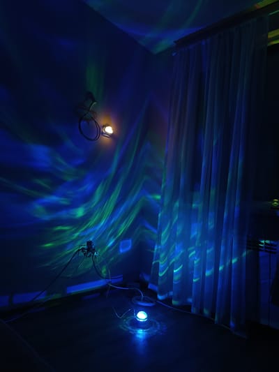 A dimly lit corner of a room is illuminated by a blue and green aurora-like light projection. The light is emanating from a round, floor-level light fixture. The projection spans the wall and partially illuminates a set of white curtains next to a radiator. Two small, warm-toned lights are mounted on the wall, further contributing to the ambiance. Electrical cords and plugs are visible. The location is not discernible as Cherkovitsa, Bulgaria.