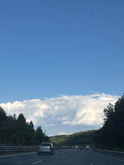 This is a vertical, eye-level, outdoor shot taken from a moving vehicle on a highway, likely in Novi Han, Bulgaria. The scene is dominated by a clear blue sky on the left and a dense line of trees bordering the highway on the right. A large, fluffy white cloud formation stretches across the middle of the frame, separating the sky from the roadside. The sun appears to be high in the sky, suggesting it is daytime, likely mid-morning or afternoon, and the weather is fair.

On the highway, several vehicles are visible, traveling in the same direction. The closest vehicle to the foreground is a silver sedan with its rear facing the camera, showing a license plate with Cyrillic characters and numbers. Further down the highway, other cars and what appear to be white vans or minibuses are also traveling. The highway itself has visible lane markings and a metal guardrail along the side. The overall impression is a typical day of travel on a highway surrounded by natural scenery.