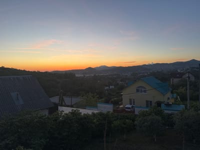 The image depicts a sunset scene over the town of Verkhnevesyoloye, Russia. The sky transitions from a pale blue at the top to vibrant orange and yellow near the horizon, with wispy pink clouds scattered across. In the foreground, there are several houses. On the left, a house with a dark, corrugated roof and a small, rectangular window is visible. To the right, a yellow house with a turquoise roof and white trim stands out, with a white car parked in front. The houses are surrounded by lush green trees and vegetation. In the background, rolling hills covered in greenery and dotted with more houses stretch towards the horizon, under the twilight sky. A power line pole is visible on the far right. The overall scene suggests a quiet residential area at the end of the day.