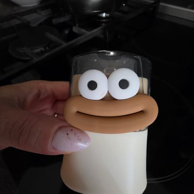 A close-up shot shows a novelty figurine with large cartoon eyes and a wide mouth. The figurine is placed on top of a white cylindrical object, possibly a container. A hand with lightly painted nails is holding the figurine. The background appears to be a dark kitchen stovetop. The image does not provide any information about the city of Peshtera, Bulgaria.