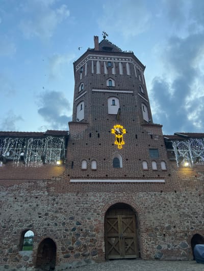 The image depicts Mir Castle in Мірскі сельскі Савет, Belarus, under an overcast sky. The central subject is a tall, octagonal tower constructed primarily of red brick. Its lower walls and the adjoining castle section are built from rough-hewn stone, transitioning to brickwork. The tower features numerous arched and rectangular windows and, midway up its facade, an illuminated yellow decorative element resembling a clock or pendulum device with hands pointing to approximately 10:10. The tower's peak includes a conical roof with a weathervane, and a significant number of dark birds are perched on its roofline and ledges, with several in flight against the sky. To the left of the tower, a section of the castle wall supports a walkway adorned with illuminated strings of cascading white lights. At ground level, a large, arched double-door wooden gate with diagonal cross bracing is centered below the tower, flanked by smaller arched and rectangular openings in the stone foundation. The ground appears to be paved with cobblestones. The illumination of the decorative lights suggests low ambient light conditions, consistent with dusk.