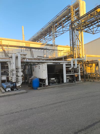 The image shows an industrial setting with large pipes, a building, and machinery under a clear blue sky. The sun is low in the sky, casting a warm golden light on the pipes and building, suggesting it is either early morning or late afternoon. The scene appears to be outdoors, with an asphalt ground surface.
Prominent objects include a large, complex network of pipes, some of which are insulated and white, while others are bare metal and yellowish, likely due to age or material. These pipes are supported by a metal framework. In the foreground, there is industrial equipment, including a large white unit with "TRANE" visible on it, suggesting it's a refrigeration or HVAC unit. Next to it is a blue plastic barrel and some smaller mechanical components. To the left, a blue motor with a pump is mounted on a concrete base.
There are no people visible in the image. The activity or event taking place is not explicitly shown, but the presence of industrial machinery and extensive piping suggests an operational or standby industrial facility. The location context provided is Dimona, Israel, which is known for its industrial activities, including a nuclear power plant and other manufacturing facilities. The overall impression is one of an industrial landscape bathed in the warm light of sunset or sunrise.