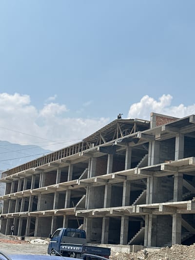 The image depicts a multi-story building under construction in Kadamjay, Kyrgyzstan. A single construction worker is visible on the roof of the building, actively working. The building is a concrete structure with visible wooden scaffolding and brickwork. A blue Hyundai truck is parked in the foreground. The environment appears to be a construction site with piles of dirt and debris. In the background, mountains and power lines are visible under a clear blue sky with scattered clouds.
