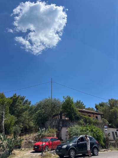 This image depicts a sunny day in Mattinata, Italy, with a clear blue sky and a large, fluffy white cloud. In the foreground, two cars are parked on a dirt road: a red Hyundai i20 on the left and a dark-colored SUV on the right. A person, dressed in a white t-shirt and shorts, is standing by the driver's side door of the SUV, appearing to be on their phone.
Behind the cars, the landscape is characterized by lush greenery, with various trees and shrubs, including pine trees, olive trees, and prickly pear cacti. A stone-built house with a tiled roof is situated on a raised terrace, surrounded by vegetation. A set of outdoor stairs leads up to the house. A utility pole with a wire stretches across the frame in the mid-ground. The overall setting suggests a rural or semi-rural area within or near Mattinata, possibly on a hillside overlooking the coast, given the typical scenery of the Gargano Peninsula.