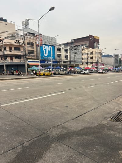 A street scene in Ban Thai–Changlon, Thailand, is depicted on an overcast day. The foreground is dominated by a paved road with white lane markings and a metal drainage grate on the right side. Along the left side of the road, a row of businesses and market stalls are visible. These include a pink and white building with balconies, a blue sign with Thai script, and various shops with colorful awnings and tents. Several vehicles are parked along the curb, including a yellow taxi, white cars, and a gray pickup truck. Pedestrians are seen walking and standing near the market stalls. A scooter is parked near one of the stalls. Overhead, numerous electrical wires crisscross the scene, supported by poles. In the background, taller buildings, including "THE VISTA HOTEL," are visible. The overall impression is a bustling urban environment with commercial activity.