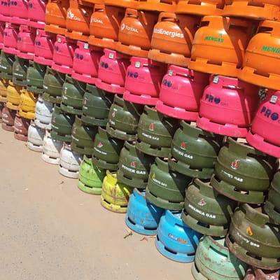 The image depicts a stack of colorful gas cylinders in what appears to be an outdoor storage area. The cylinders are stacked in rows, with different brands such as "TOP Gas", "TOTAL", "TotalEnergies", "MENGAS", "PRO Gas", "POWER Gas", "KENTO Gas", "MWANGA Gas", and "Lake Gas" being visible. The cylinder colors vary across rows, including orange, pink, green, blue, yellow, white, and brown. The cylinders are stacked on a concrete or asphalt surface. Based on the OCR and brand names, it's likely the image was taken in an African country. The city of Sheywe ward in Kenya is not explicitly visible in the image, but it is implied as the setting of the image. There are no people visible in the image. The cylinders appear to be in varying states of wear and tear.