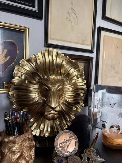 The image depicts a desk with several decorative items and office supplies, likely in a home or office setting. The primary focal point is a large, ornate gold lion head sculpture with a detailed mane. To the left of the lion head is a framed piece of art with a dark background and a gold design. In front of the lion head, a pen holder shaped like a gold lion's head is filled with various pens and writing utensils. A circular bronze medal with an image of a dog and text reading "LEADER DOGS FOR THE BLIND" is positioned in front of the pen holder. To the right of the lion head sculpture is a clear acrylic framed clock with Roman numerals. A small wooden base holds an award with a star and circle design. The background features framed artwork, including sketches. The overall impression is one of a curated display of personal or professional memorabilia. The location is identified as Sun City Center, United States of America.
