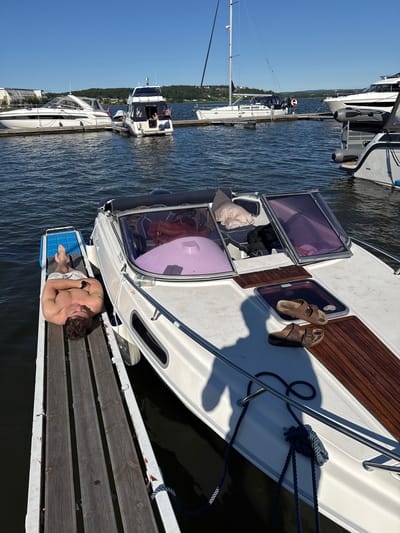 A shirtless man is lying on a wooden dock next to a white boat in Tønsberg, Norway. The man is wearing patterned shorts and has his arms crossed over his chest. The boat has a purple tinted windshield and a wooden deck. A pair of brown sandals are placed on the boat's deck. Other boats and a sailboat are docked in the background. The sky is clear and blue.
