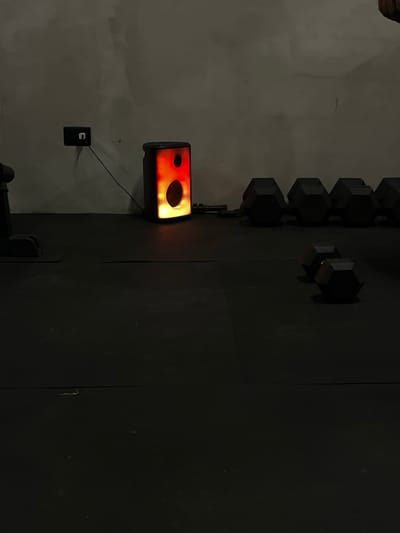 An indoor scene at a gym in an unknown city in Mexico shows a dark, textured concrete wall behind a dark floor. To the left, a black electrical outlet is mounted on the wall with a thin black wire extending to a rectangular speaker with a lit interior emitting orange and yellow light. In the background, a row of black, hexagonal dumbbells is visible on the floor, with two more dumbbells in the foreground.