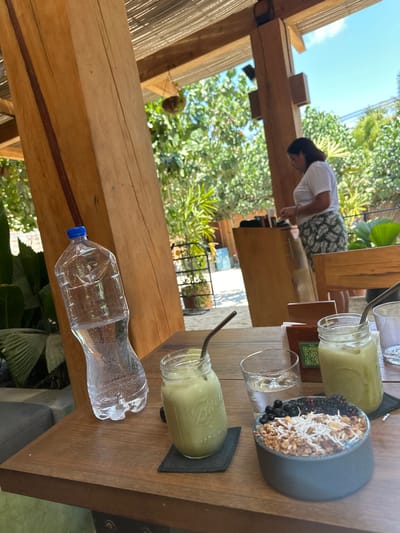 The image shows a wooden table with a bottle of water, two green smoothie-like drinks in jars with straws, and a bowl of what appears to be granola, coconut flakes, and blackberries on top. A woman in a white shirt and patterned shorts stands beyond the table. The setting seems to be outdoors, with trees and sky visible in the background. The location is Puerto Escondido, Mexico.