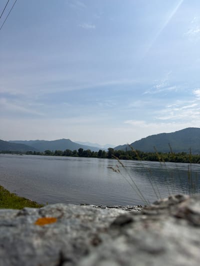 A wide shot captures a serene natural landscape in the vicinity of the village of Sizaya, Russia. The foreground is dominated by a rough, grey stone surface, out of focus, with a single, vibrant orange leaf resting upon it. Beyond the stone, a vast, calm river flows, its surface rippled by a gentle breeze. The river is bordered by lush green vegetation and trees along its banks. In the mid-ground, a line of dense trees forms a natural boundary before the land begins to rise into a series of rolling, tree-covered hills and mountains that recede into the distance, softened by atmospheric haze. The sky above is a pale blue, sparsely dotted with wispy white clouds, and a faint sunbeam streaks across the upper right quadrant of the frame. The overall scene evokes a sense of tranquility and natural beauty.