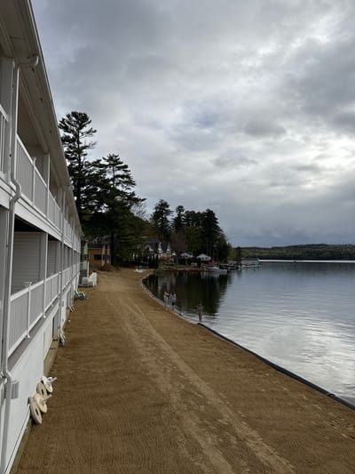 The image shows a lakeside scene in Laconia, United States of America. From the perspective of a white structure with latticed patterns, a sandy beach stretches to the right, meeting the calm water of a lake. In the distance across the water, forested land is visible. The sky above is overcast with gray clouds. There are a few visible houses amongst trees near the shoreline in the mid-ground. A retaining wall or edging separates the beach from the water. There are some objects that appear to be large spools on the roof, as well as some PVC pipes.