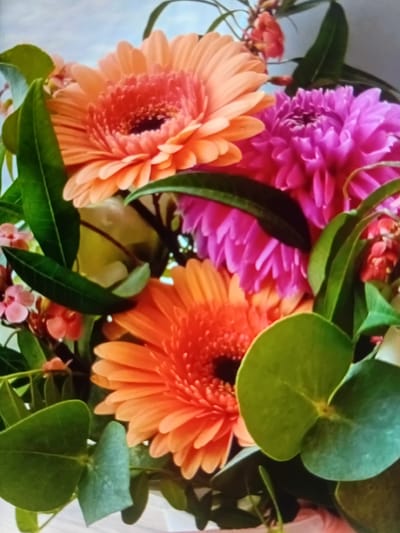 A close-up shot of a bouquet of flowers, featuring gerbera daisies in shades of orange and pink, along with other blossoms and greenery. The bouquet is arranged in a way that fills the frame, with the flowers being the primary focus. The background is softly blurred, suggesting an indoor setting or a shallow depth of field. There are no people or specific identifiable landmarks that would indicate a location in Verkhniachka, Ukraine, or any other city. The image is a still life composition of flowers.