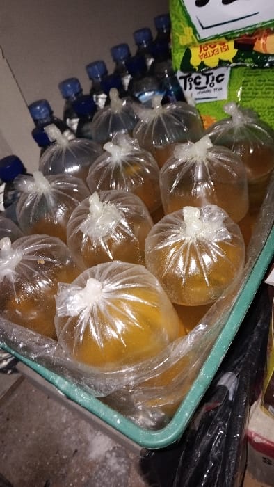 A green plastic bin contains numerous clear plastic bags, each filled with a yellow liquid. The bags are clustered together in the bin and are partially obscured by a clear plastic bag extending from the bin. Some of the bags are tied at the top. The bin is on a shelf, and there are numerous blue water bottles behind it. The bin is located in a room that appears to be a store in Kecamatan Panjalu, Indonesia, based on the provided coordinates.