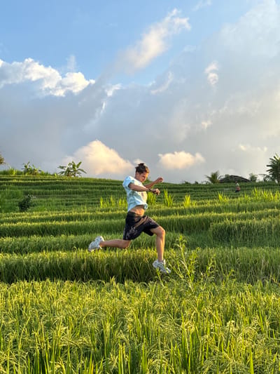 A man is captured mid-air, positioned at an approximate 45-degree angle against a backdrop of a rice field. The location is Padangbulia, Indonesia. The man wears light-colored t-shirt, dark shorts, and light-colored sneakers. He is in the middle of a backflip. The rice field features rows of green stalks. The sky behind the rice field is filled with white clouds and a gradient of light blue to a near-white sky.