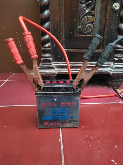 A black "GS PREMIUM GM5Z-3B 12V 5Ah (10HR)" automotive battery is centrally positioned on a reddish-brown tiled floor with white grout lines. A red-handled jumper cable clamp is connected to the battery's left terminal, with its red insulated cable extending upwards and curving behind the battery. A black-handled jumper cable clamp is connected to the battery's right terminal, with its black insulated cable extending horizontally to the right, out of frame. Both the battery and the floor exhibit dust and minor debris. The background features a dark, ornate wooden cabinet or furniture piece, characterized by twisted vertical columns and carved decorative elements. The wooden background also shows accumulated dust. The image contains no discernible information to confirm the location as Mornington, Australia.