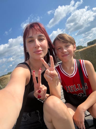 Two individuals, appearing to be a woman and a child, are captured in a selfie taken outdoors. The woman, with reddish-brown hair, is in the foreground, her face partially visible. She is wearing a black tank top and has red nail polish on her fingernails. She is making a peace sign with her right hand. The child, with light brown hair, is behind her, also making a peace sign with their left hand. They are wearing a red basketball jersey with white trim and black lettering that reads "CHICAGO BULLS". They are smiling at the camera. The background shows a bright blue sky with scattered white clouds, suggesting it is daytime with fair weather. Hints of hay bales and trees are visible, and the people are seated on what appears to be a wooden surface, possibly a dock or deck. The overall mood appears to be relaxed and cheerful, consistent with a casual outing.