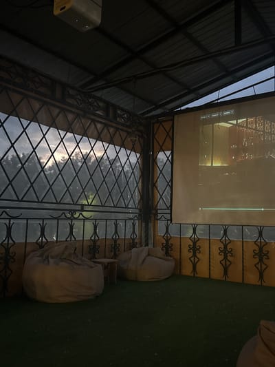 The image captures an outdoor seating area, possibly a patio or balcony, with a projector screen set up. The screen displays what appears to be a menu or interface with text elements such as "Co-Op I", "Common I", and "SoftBoss". A projector is mounted on the left side of the image, pointing towards the screen.
The setting includes a metal roof structure over part of the area, providing shade. Decorative wrought iron fencing is visible, with a diamond pattern. Outside the fence, there's a reflection of the sky with clouds, suggesting it might be daytime or dusk. Two light-colored beanbag chairs are placed on the ground, indicating a casual seating arrangement for viewing the projector. The ground appears to be covered in a dark green, textured material, possibly artificial turf or painted concrete.
The overall atmosphere seems relaxed and geared towards entertainment, like an outdoor movie night or a sports viewing event. The lighting suggests it's late afternoon or early evening. The location is identified as Cherkessk, Russia, which provides geographical context. No people are visible in the frame.