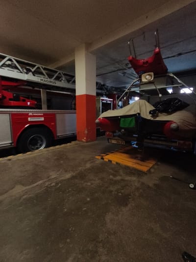 The image shows a fire truck inside a garage. The fire truck is red and silver, with the brand name "MAGIRUS" visible on the side. The garage appears to be dimly lit, with exposed ceiling beams and some structural supports. In the foreground, the concrete floor is visible, with some yellow and black hazard markings on the ground. In the background, a second vehicle, possibly a trailer, is covered with a tarp. The scene appears to be in a storage or maintenance area for emergency vehicles. There is no specific indication of the location in the city of Tukums, Latvia, within the image itself.