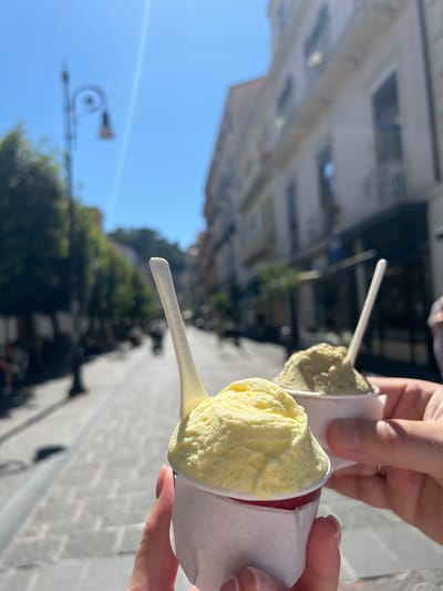 In the foreground, two hands hold white paper cups of gelato. The cup in the left hand contains pale yellow gelato, possibly lemon or vanilla, with a red base. The cup in the right hand holds a lighter green gelato, possibly pistachio. Both cups have white plastic spoons. The background is blurred and depicts a sunlit, cobbled street lined with trees and buildings, characteristic of Sorrento, Italy. A traditional street lamp is visible on the left. The sky is a clear blue. Several blurry figures are walking along the street.
