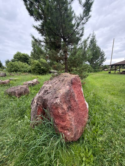 A large red boulder is in the foreground of a grassy field. A pine tree is behind the boulder, with other trees and bushes in the background. A gazebo is visible in the distance, as is a utility pole. The sky is overcast.