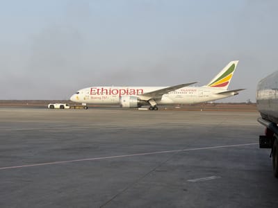 The image depicts an Ethiopian Airlines Boeing 787 Dreamliner on an airport tarmac. The aircraft is painted white with the word "Ethiopian" in red along the fuselage. The tail fin has the colors green, yellow, and red. The registration number "ET-AOP" is visible near the tail. In the background, there is a small vehicle, and to the right, part of a fuel truck is visible. The sky is overcast, suggesting hazy conditions. The scene appears to be on the ground, as there are ground crew vehicles visible. The image was taken in the city of Communauté rurale de Diass, Senegal.