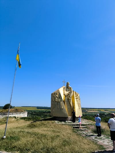 The image depicts an outdoor scene on a clear, sunny day at the Monument to the Khotyn Battle, situated on a hill overlooking the Khotyn Fortress and the Dniester River valley in Khotyn, Chernivtsi Oblast, Ukraine.

In the foreground, an uneven, dry grassy slope leads up to a dirt path with stone steps. To the left, a tall metal flagpole bears a Ukrainian flag (blue over yellow), hanging loosely due to minimal wind. The flag pole is positioned on a raised earthwork, likely part of the historical fortifications.

The central subject is a large, multi-faceted golden monument, likely constructed from a metal or stone material with a gold finish. The monument features a prominent, stylized figure, resembling a religious leader or saint, adorned with a cross on top, and flanked by bas-relief panels depicting multiple other figures. It stands on a stone base with several steps leading up to it.

Four individuals are present near the monument. An adult female dressed in dark clothing stands at the immediate base of the golden monument. A young child, wearing a pink hat and light-colored attire, descends the stone steps from the monument. To the right of the path, an adult male in a light blue shirt and dark pants holds a camera to his eye, appearing to photograph the scene. Another adult male, wearing a white t-shirt and dark shorts, stands slightly behind and to the right of the camera-wielding individual, with his back partially to the viewer.

In the mid-ground and background, remnants of stone fortifications or ruins are visible to the left of the monument. Beyond these structures and the monument, a vast landscape unfolds, comprising a valley with cultivated fields (some appearing green, others golden or brown) and scattered clusters of trees, extending to a distant horizon under a cloudless, bright blue sky.
