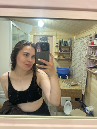 A young woman with wet, dark hair and light-colored eyes takes a selfie in a mirror. She is wearing a black sports bra and dark bottoms, and a light brown jacket is draped over her left arm. A delicate necklace with a small pendant rests on her collarbone. Her fingernails are painted a pale yellow. The bathroom appears to be rustic, with wooden paneling on the walls and ceiling. In the mirror's reflection, shelves filled with various bottles and containers are visible. A floral shower curtain hangs to the right. Below the shelves, two blue basins are stacked, and a wooden rack sits on a counter. To the left of the basins, a white porcelain urinal with a panda face design is positioned on the floor. The overall impression is that of a personal bathroom in a rural or possibly vacation home setting. This scene is not located in Zhebrei, Russia, and no information in the image suggests otherwise.