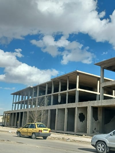 The image depicts an urban street scene in Al-Qadam Municipality, Syria, under a blue sky with scattered white cumulus clouds. A large, multi-story concrete skeletal structure, approximately four stories high, dominates the background, appearing either under construction or partially damaged. The building's concrete framework of columns and slabs is visible, with some lower sections featuring incomplete brick infill walls, one of which exhibits a large, irregular circular void. A bare, deciduous tree stands in front of the building's lower section. On the paved road in the foreground, a yellow four-door sedan with a roof-mounted sign is positioned on the left, facing away from the viewer, showing a visible dent on its rear driver-side door. The front right wheel and bumper section of a silver SUV are partially visible in the bottom right corner. The road surface and adjacent dirt shoulder show scattered debris. No individuals are present in the scene.