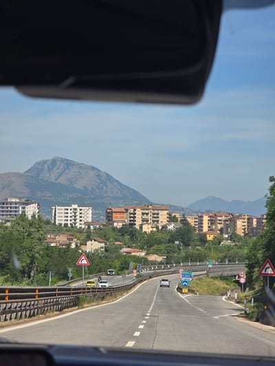 A road curves through a town with mountains in the background. The road is made of asphalt and has white dashed lines marking the lanes. There are several cars on the road, including a silver SUV, a yellow car, and a white car. There are also several buildings visible in the background, including apartment buildings and houses. The mountains in the background are covered in trees. The sky is blue and there are a few clouds. The image appears to be taken from inside a car, looking out through the windshield. There are traffic signs visible along the side of the road, including a speed limit sign of 40 km/h and a curve warning sign. One sign indicates directions to "Benevento est", "Avellino", and "Caserta".