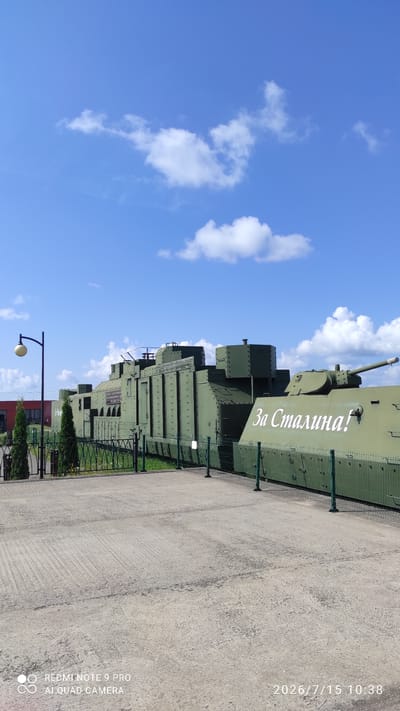A large, green, armored train car with "За Сталина!" painted on its side is on display outdoors under a partly cloudy blue sky. The train car is long and appears to be made of metal plates bolted together. It has several turrets and appears to be a historical exhibit. In the foreground, there is a paved area with a textured surface. Behind the train car, there is a green metal fence and some trees. To the left, there is a lamppost. The overall scene suggests an outdoor museum or historical site.