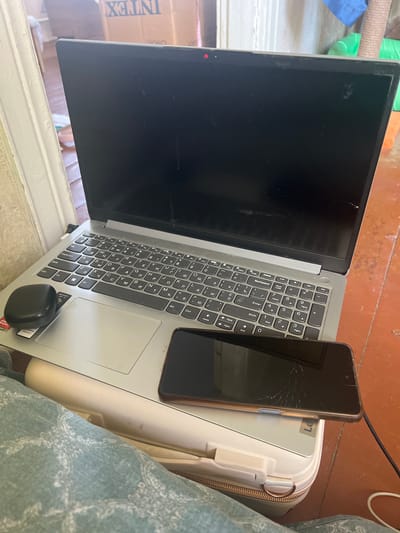 An open laptop is on a white briefcase. A smartphone with a cracked screen lies on top of the laptop. A black wireless mouse is on the left side of the laptop's touchpad. The background shows boxes and a wooden floor. The image appears to be taken indoors. The user states this image is from Buzova, Ukraine.
