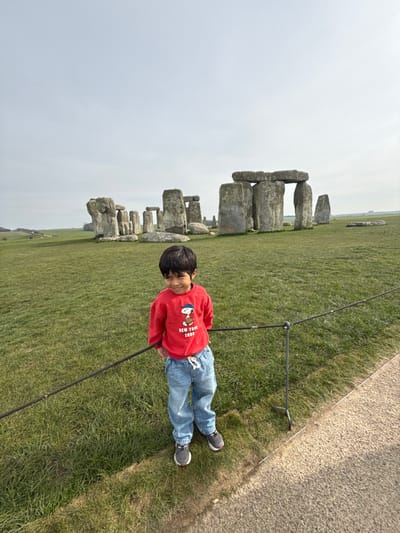 A young boy stands smiling in the foreground, positioned behind a dark rope barrier that separates a grassy field from a paved pathway. The boy is wearing a red long-sleeved shirt with a Snoopy graphic and "NEW YORK 2000" text, light blue jeans, and dark athletic shoes. His hands are clasped behind his back. In the background, the prehistoric monument of Stonehenge is visible, consisting of large, grey standing stones and trilithons arranged in a circular formation on a vast green field. The sky is overcast. The scene is located at Stonehenge, near Amesbury, Wiltshire, United Kingdom.
