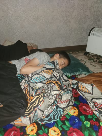 A young boy sleeps on a bed, his head resting on a teal pillow. He is covered by a blanket with a colorful floral pattern, featuring red, blue, yellow, and green flowers. Another blanket with a geometric print in shades of gray, blue, and orange is also visible. The boy is wearing a light gray t-shirt. To the left, a person wearing black clothing is partially visible, also appearing to be resting. The background shows a wall with patterned wallpaper and a white radiator on the right side of the image. The scene appears to be indoors, likely a bedroom in a residential setting in the city of Yefremov, Russia.