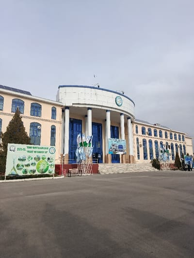 A large, multi-story building dominates the frame, its facade featuring a rounded central section supported by white columns and blue-trimmed windows. Signs and flags are positioned in front of the building, adding visual interest. The paved area in the foreground stretches to the building, with trees visible on either side, all set under an overcast sky.