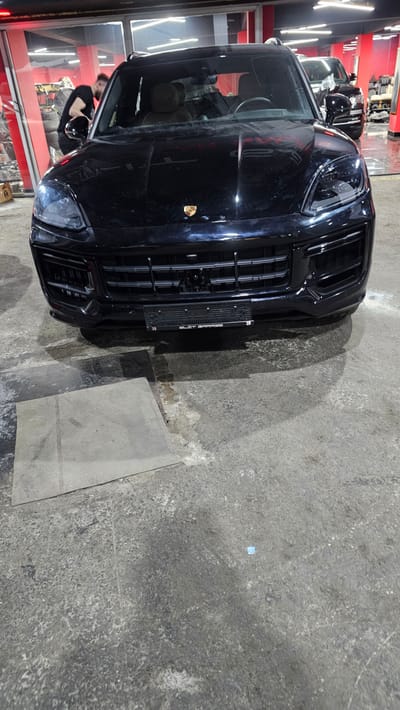 A black Porsche Cayenne SUV is parked on a concrete floor inside what appears to be a car service or detailing center. The front of the vehicle is prominently displayed, showing its headlights, grille, and license plate. A person, wearing a black t-shirt, is visible in the driver's side window, leaning in towards the car. In the background, through glass doors, other vehicles and car detailing equipment can be seen. The overall environment suggests an indoor automotive facility. The image is taken from a slightly low angle, looking up at the front of the SUV.