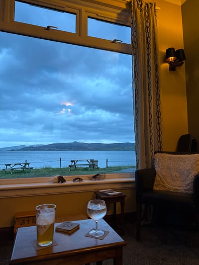 This image captures an interior view looking out a large window towards a body of water in Sconser, United Kingdom. The multi-pane window features white frames, with a brass-colored handle visible on the upper left sash.

The exterior scene, viewed through the window, consists of a calm, dark blue body of water in the midground, separating a strip of green grass in the foreground from a distant landmass of rolling hills and mountains under a heavily overcast, blue-grey sky. Two wooden picnic tables are positioned on the grass directly outside the window, along with a wooden fence post. Two bright, rectangular reflections, likely from indoor light sources, are prominent in the upper center of the main window pane.

The interior of the room features a wall painted in an olive-green tone on the far right, transitioning to a warmer yellow/ochre hue around the window and above the wall sconce. Beige curtains with a repeating dark brown chain-link pattern are drawn to the right of the window. A wall-mounted sconce with two black, conical lampshades emitting a warm orange light is affixed to the right wall. In the lower right, a dark brown armchair is visible, adorned with a light-colored, fluffy cushion. The floor is covered with dark grey carpeting.

In the immediate foreground, a small, dark wooden table holds a pint glass, approximately two-thirds full of an amber liquid with a white foam head, resting on a square coaster bearing the text "SKYE ALE." Alongside it, a stemmed glass, half-full of clear liquid, rests on a round coaster. To the right of this table, a smaller, darker wooden side table holds a book or magazine. A small, ornate wooden box is partially visible beneath the foreground table on the left. The windowsill itself is adorned with several decorative items, including a large conch shell, a dark hermit crab figurine, a dark crab figurine, and a small wire basket. No individuals are present in the scene.