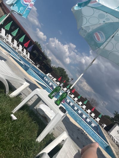 The image depicts a sunny day at an outdoor swimming pool, possibly a resort or a private establishment, in Manolsko Konare, Bulgaria. The scene is set with a long, rectangular swimming pool filled with clear blue water, lined with white lounge chairs and small, colorful umbrellas (red, green, and blue). The poolside is paved with grey concrete.

In the foreground, two green glass bottles and a white plastic chair are visible. The chairs are tilted, suggesting they are unoccupied. The sky is a mix of bright blue with fluffy white clouds and darker, more ominous storm clouds, hinting at a dynamic weather situation. The overall atmosphere seems relaxed, typical of a summer day by the pool, though the storm clouds might introduce a slight sense of unease.

A prominent detail is a large, partially visible blue and white umbrella with a repeating circular pattern and the "Nestlé" logo. This suggests a branding or sponsorship at the location. The image is taken from a low angle, possibly by someone lying on a lounge chair, as indicated by the presence of a foot in the lower right corner. The sun casts distinct shadows, confirming it is daytime, likely late morning or afternoon. The lush green grass bordering the paved area adds to the outdoor setting. There is no clear activity or event taking place, other than the implied leisure use of the pool area.