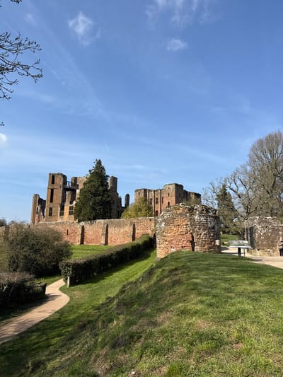 The image displays an exterior view of Kenilworth Castle ruins in Kenilworth, Warwickshire, United Kingdom, under a clear blue sky with scattered wispy white clouds.
In the foreground, a winding dirt path is visible on the left, bordered by low, dense green shrubs and ascending green grass. To the right, a grassy mound leads to a circular, reddish-brown stone structure, partially covered in white lichen. Adjacent to this, a gravel path features a metal information stand with an interpretive panel. A single individual, clad in dark clothing, is partially visible on the far right, standing near the gravel path.
The midground is dominated by a long, low wall constructed from reddish-brown stone, topped with a neatly trimmed green hedge. Below this, another manicured hedge follows the contour of a grassy slope.
The background prominently showcases the extensive, multi-story ruins of Kenilworth Castle, characterized by their reddish-brown stone construction. Visible window openings and fragmented wall sections form the dilapidated structures. A tall, dark green coniferous tree stands centrally within the castle ruins, while several large, bare deciduous trees are situated on the right periphery, behind additional castle segments. The overall environment suggests a historical site on a bright, possibly early spring, day.