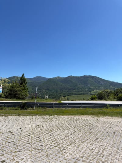 The image shows a view from a paved rest area or parking lot adjacent to a road, likely a highway. The paving material is permeable concrete. Beyond the road is a green, mountainous landscape under a clear blue sky. There's a guardrail separating the road from the parking area. Vegetation is growing near the road's edge. The location is somewhere in the city of Bunovo, Bulgaria. There are road signs. There are no people visible.